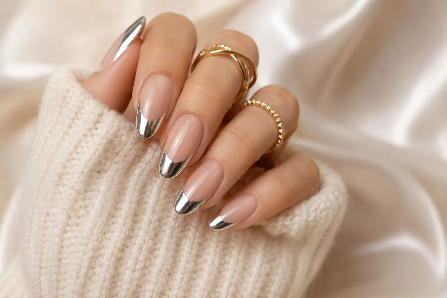 Reflective chrome French tips on almond-shaped nails with gold rings and a cream knit sleeve.